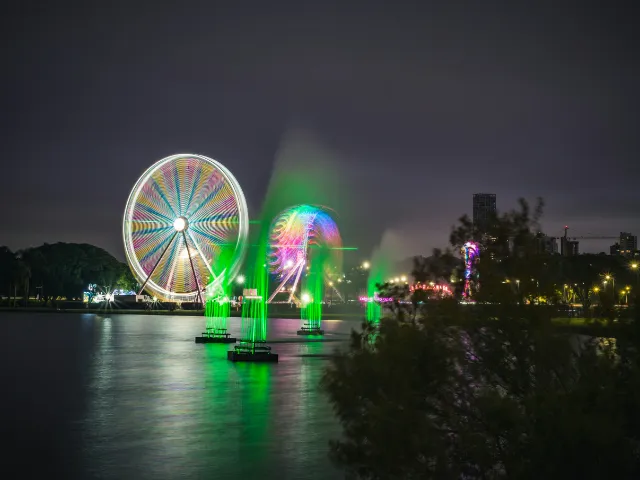 A ferris wheel and green fountains lit up at night over a lake