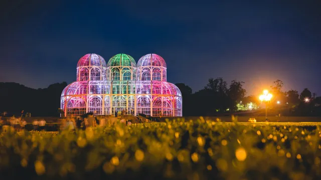 A glass greenhouse lit in pink and green at night, behind a field of flowers